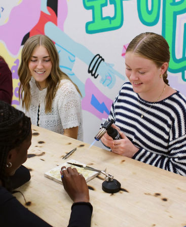Three people at a colorful art studio craft workshop; one uses a handheld torch to heat a small jewelry project on a fireproof tile while others watch and assist.