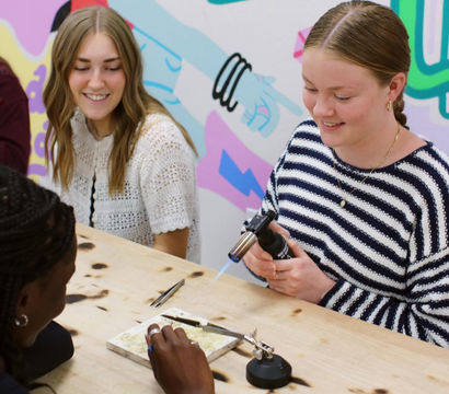 Three people at a colorful art studio craft workshop; one uses a handheld torch to heat a small jewelry project on a fireproof tile while others watch and assist.