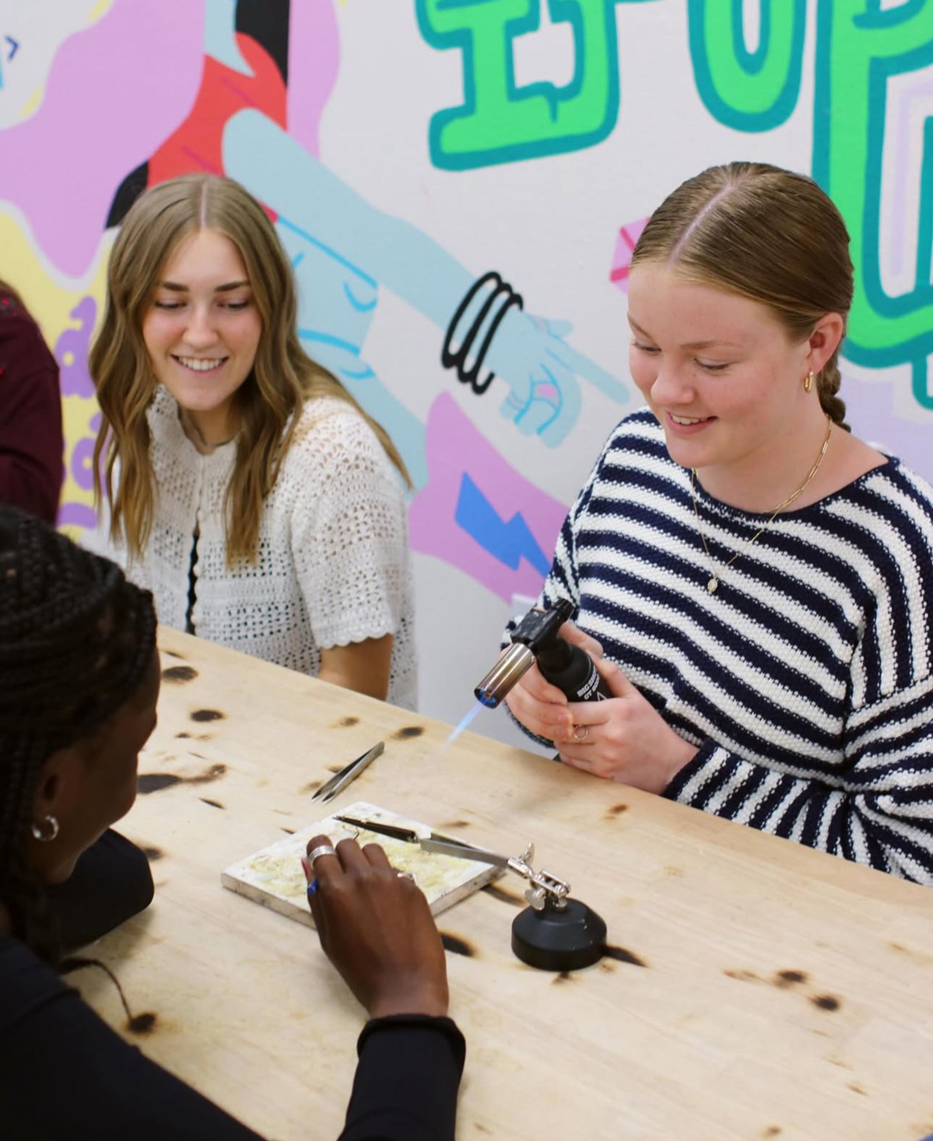 Three people at a colorful art studio craft workshop; one uses a handheld torch to heat a small jewelry project on a fireproof tile while others watch and assist.