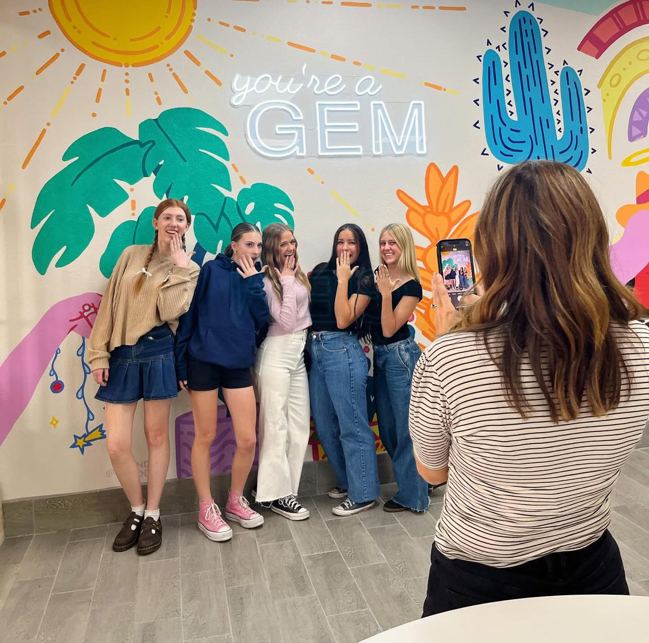 Five friends strike playful ring-showing poses while someone photographs them on a smartphone in front of a colorful indoor mural reading “you're a GEM” with sun, palm leaves and a cactus.