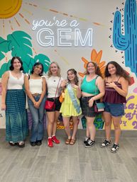 Six friends posing indoors in front of a colorful desert mural and neon 'you're a GEM' sign; central woman in a lime dress and sash, group in casual summer outfits
