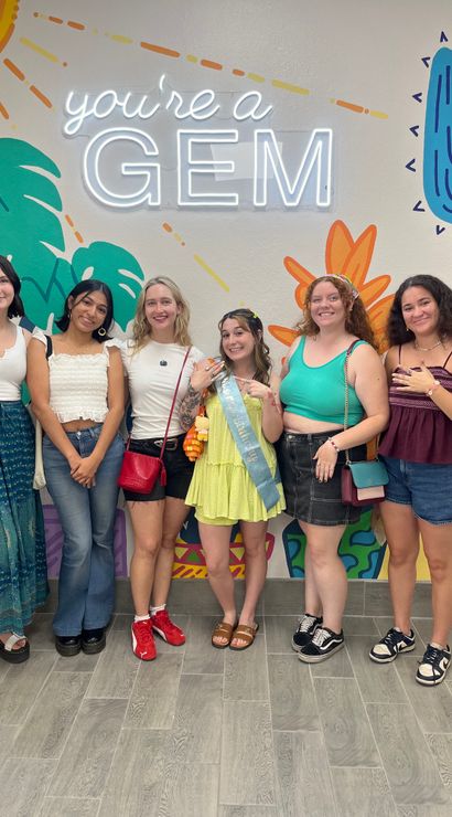 Six friends posing indoors in front of a colorful desert mural and neon 'you're a GEM' sign; central woman in a lime dress and sash, group in casual summer outfits