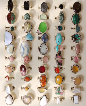 Colorful display of handmade silver gemstone rings on a cream tray — turquoise, malachite, agate, jasper, opal and cameo styles arranged in neat rows.