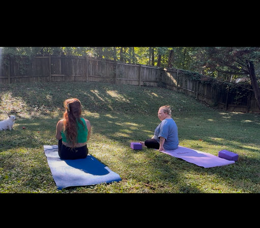 Two people enjoying backyard yoga on blue and purple mats in a sunny, fenced grassy yard with yoga blocks and a small white dog nearby.