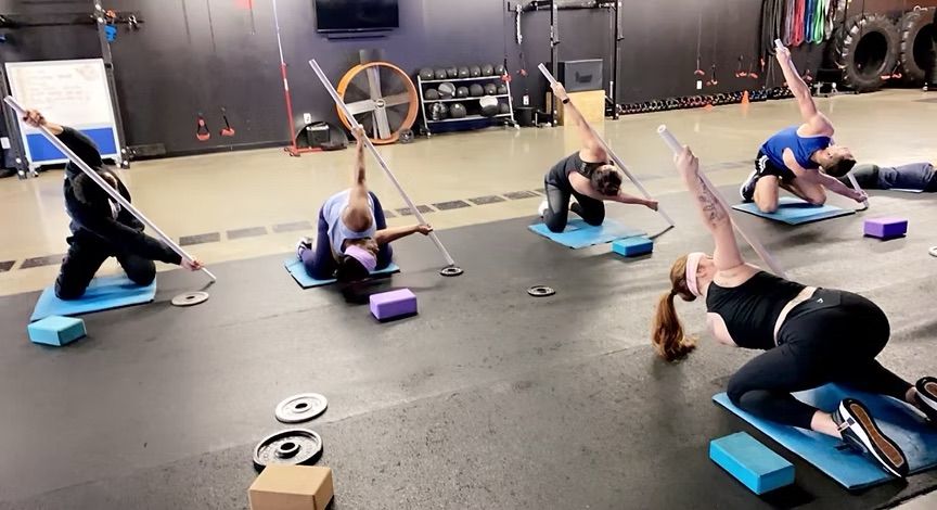 Group fitness class in a gym performing seated thoracic rotation mobility with dowel rods on yoga mats and foam blocks, surrounded by weight plates and large tires.