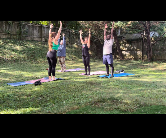 Four people on yoga mats doing a standing arm‑raise in a sunny backyard lawn with trees and a wooden fence — outdoor group yoga/stretch session.