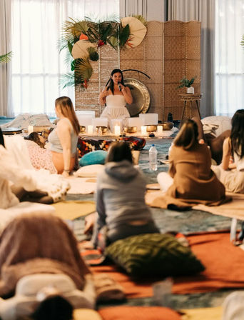 Group sound bath meditation in an indoor wellness retreat — instructor in white with microphone by a gong and singing bowls, attendees seated on cushions amid candles and plants.