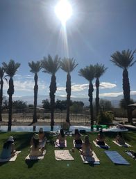 Sun-drenched poolside yoga on a green lawn with a row of palm trees and desert mountains beyond under a bright midday sun.