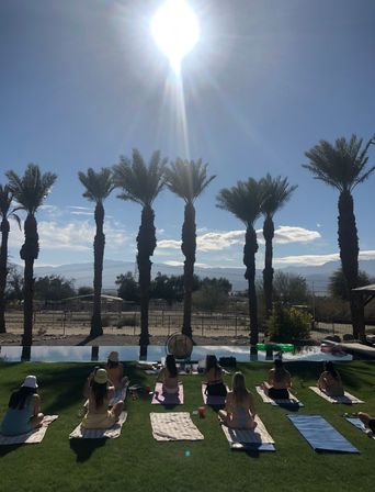 Sun-drenched poolside yoga on a green lawn with a row of palm trees and desert mountains beyond under a bright midday sun.