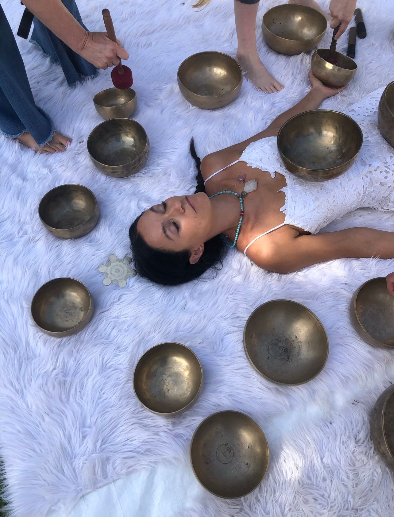 Woman lying on a fluffy white rug surrounded by bronze singing bowls while practitioners use mallets in an outdoor sound-healing session, wearing a white lace dress and crystal necklace.