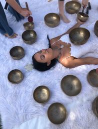 Woman lying on a fluffy white rug surrounded by bronze singing bowls while practitioners use mallets in an outdoor sound-healing session, wearing a white lace dress and crystal necklace.