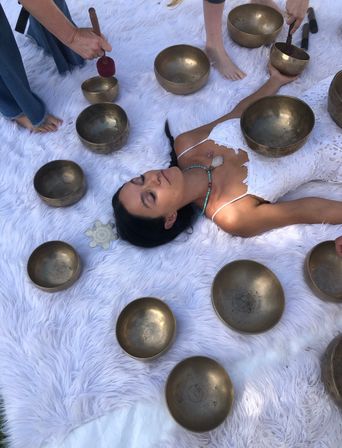 Woman lying on a fluffy white rug surrounded by bronze singing bowls while practitioners use mallets in an outdoor sound-healing session, wearing a white lace dress and crystal necklace.