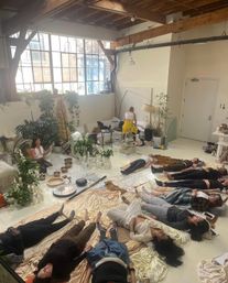 Sunlit urban loft studio with exposed wooden beams and large windows where people lie on blankets for a guided sound-bath meditation, instructor seated among singing bowls, gongs, candles and lush plants — a relaxed wellness session.