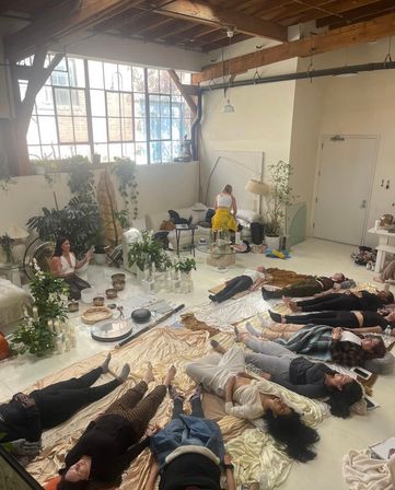 Sunlit urban loft studio with exposed wooden beams and large windows where people lie on blankets for a guided sound-bath meditation, instructor seated among singing bowls, gongs, candles and lush plants — a relaxed wellness session.