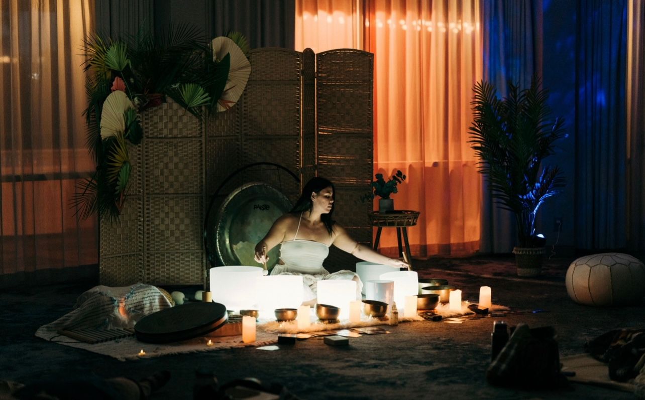Person leading a sound bath, sitting among glowing crystal singing bowls, candles, brass bowls and a gong in a dim indoor meditation space with warm orange and blue curtain lighting and potted plants.