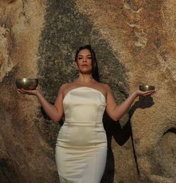 Woman in a strapless cream dress standing against a rugged rock formation, arms outstretched holding two brass singing bowls in a balanced, meditative pose bathed in warm golden light.