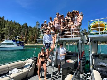 Group of friends posing on a two-level pontoon boat at a turquoise mountain lake with pine-tree shoreline and clear blue summer sky