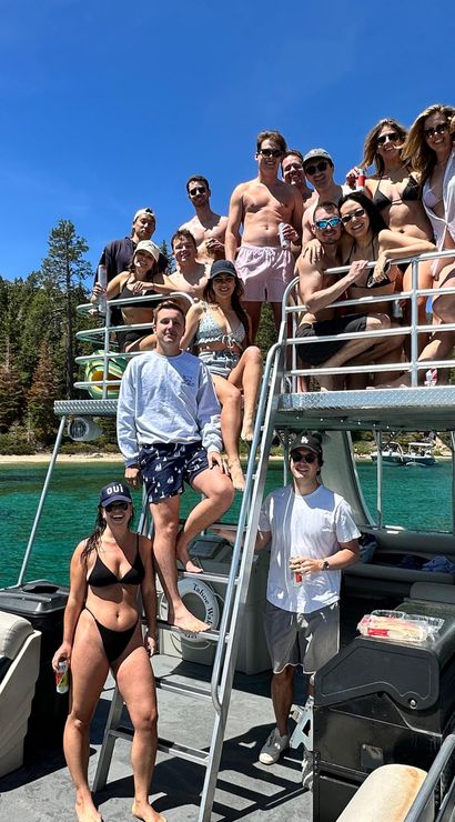 Group of friends posing on a two-level pontoon boat at a turquoise mountain lake with pine-tree shoreline and clear blue summer sky