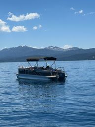 Pontoon boat bobbing on a calm blue mountain lake under a bright sky with distant snow-dusted peaks and fluffy clouds