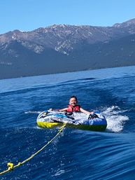 Smiling child in a red life jacket tubing on a colorful inflatable, being towed across a deep blue alpine lake with forested shoreline and snow-dusted mountains under a clear sky.
