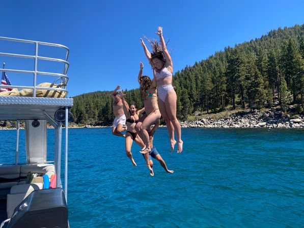 Four people mid-air jumping off a boat into a clear blue alpine lake, framed by a boat railing, pine-covered hillside and rocky shoreline under a bright sunny sky.