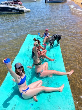 Group of women relaxing on a turquoise floating mat at a sunny lake beach, raising drinks while a spotted dog explores nearby and boats float in the background