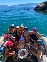 Friends in colorful wigs and tie-dye bucket hats posing on a sunlit boat over crystal-clear Lake Tahoe waters with rocky shoreline and distant mountains.