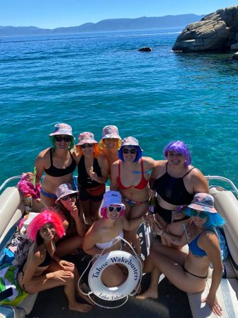 Friends in colorful wigs and tie-dye bucket hats posing on a sunlit boat over crystal-clear Lake Tahoe waters with rocky shoreline and distant mountains.