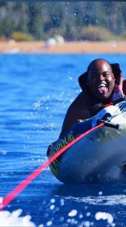 Man lake tubing on an inflatable towable, grinning with tongue out and life vest on, pulled by a red rope across blue water with splashing waves and a sandy tree-lined shore in the background.