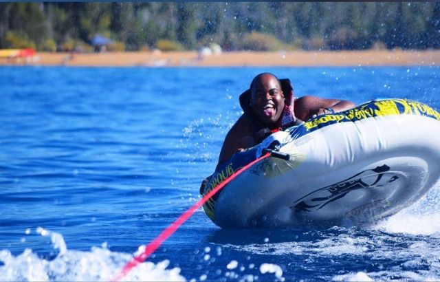 Man lake tubing on an inflatable towable, grinning with tongue out and life vest on, pulled by a red rope across blue water with splashing waves and a sandy tree-lined shore in the background.