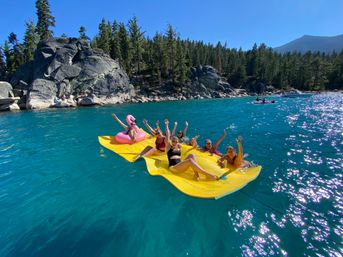 Friends waving on a yellow inflatable mat and pink flamingo float in a bright turquoise alpine lake with granite boulders, pine-forested shoreline and clear blue sky