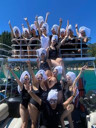Fun group of friends in matching black swimsuits and white sunglasses with towel turbans, cheering on a pontoon boat over a turquoise lake with pine-covered shoreline and clear blue summer sky.