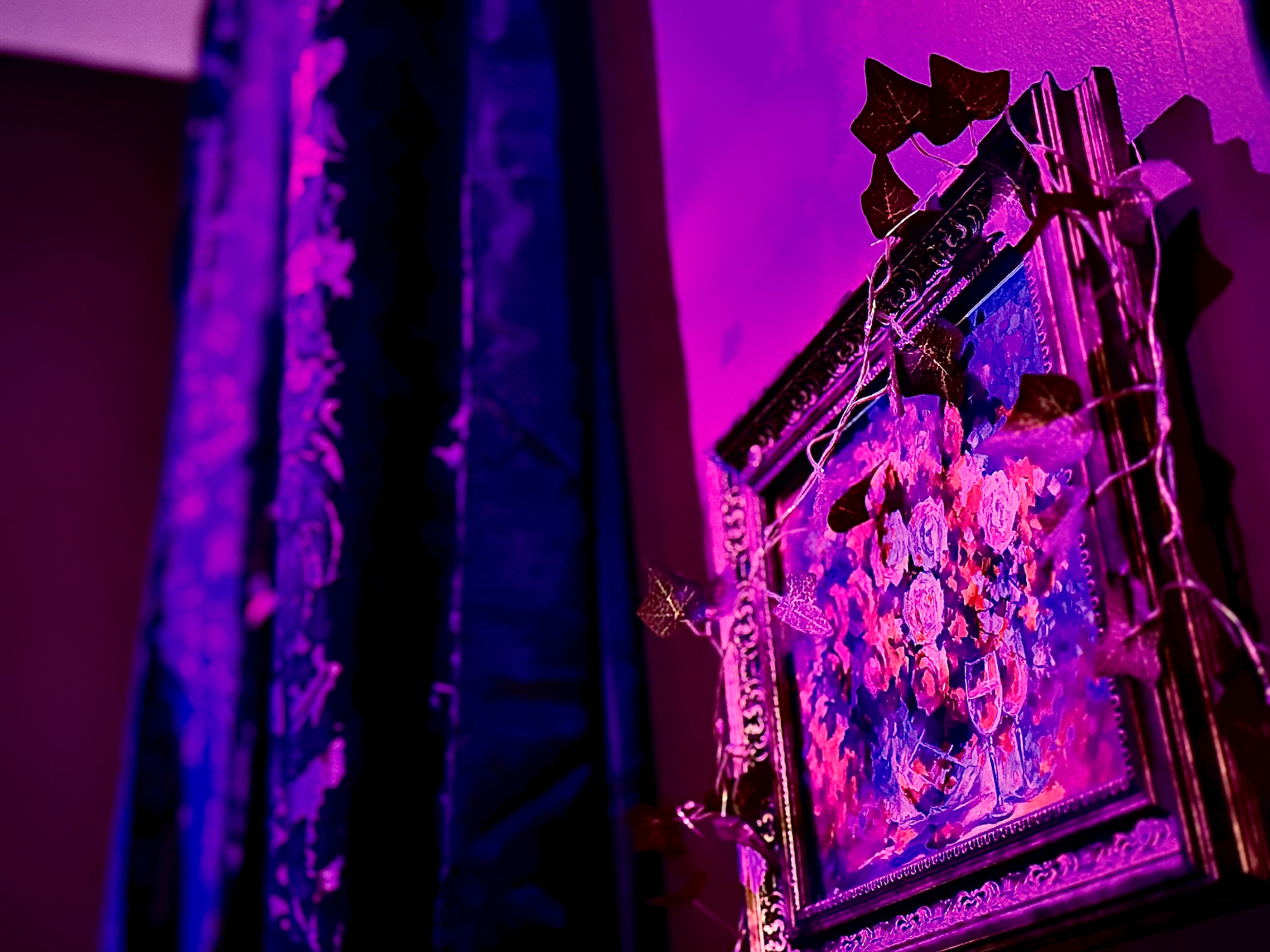 Vibrant magenta-lit corner featuring an ornate framed floral painting wrapped in leaf string lights beside dark patterned curtains — moody indoor wall art and decor.