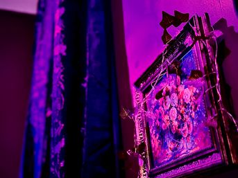 Vibrant magenta-lit corner featuring an ornate framed floral painting wrapped in leaf string lights beside dark patterned curtains — moody indoor wall art and decor.