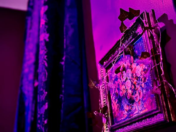 Vibrant magenta-lit corner featuring an ornate framed floral painting wrapped in leaf string lights beside dark patterned curtains — moody indoor wall art and decor.