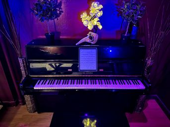 Black upright piano in a cozy home music room bathed in moody purple-blue light, sheet music on the stand, an ornate decorative mask and bright yellow faux flowers on top, flanked by potted plants and tall wicker vases.