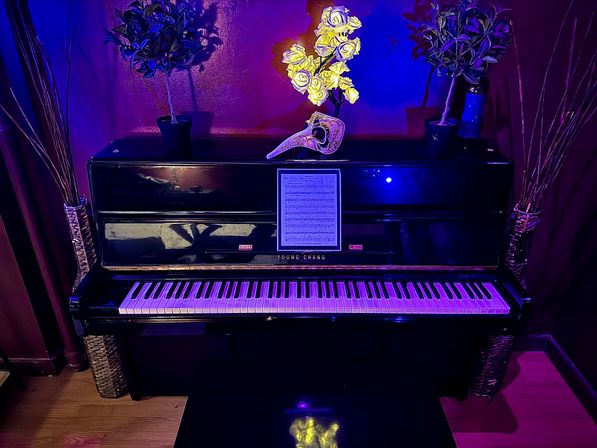 Black upright piano in a cozy home music room bathed in moody purple-blue light, sheet music on the stand, an ornate decorative mask and bright yellow faux flowers on top, flanked by potted plants and tall wicker vases.