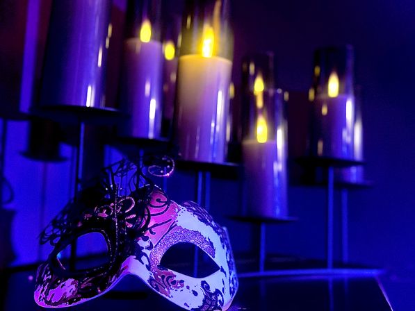 Ornate masquerade mask on a tabletop beside glowing pillar candles in glass holders, moody purple-blue lighting — indoor party or theatrical decor.