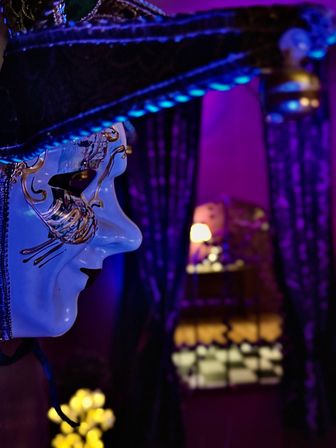 Close-up profile of an ornate white Venetian carnival mask with gold filigree and wide hat in a purple-lit room, blurred lamp, curtains, and warm bokeh lights in the background