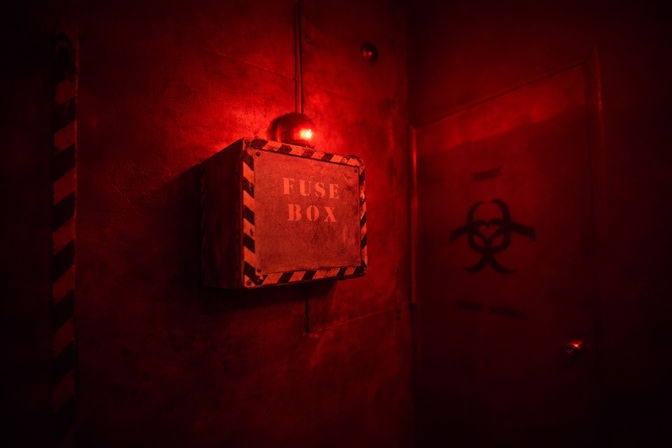Red emergency-lit industrial hallway with a wall-mounted metal fuse box labeled 'FUSE BOX' with hazard stripes and a glowing red alarm, next to a door marked with a biohazard symbol.