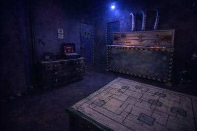 Moody industrial escape-room set with blue overhead light, vintage control panels on a worn chest, a hazard-striped master console against the wall, and a schematic-covered worktable in the foreground.