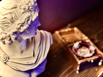 Close-up of a white classical-style marble bust on a wooden table beside a vintage wooden box and ashtray, moody purple lighting, home decor vignette.
