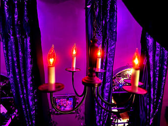 Ornate metal chandelier with four glowing candle-shaped bulbs casting warm red-orange light on purple-lit velvet drapes and a decorative mirror in a moody indoor setting.