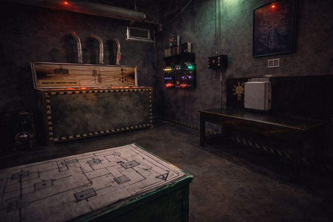 Moody dimly lit industrial basement control room with vintage electrical panels and colored indicator lights, a metal chest with caution trim, a wooden bench with a metal case, and a blueprint-topped table in the foreground.