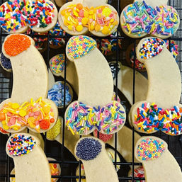 Playful novelty penis-shaped sugar cookies topped with assorted colorful sprinkles and nonpareils, arranged on a black cooling rack.