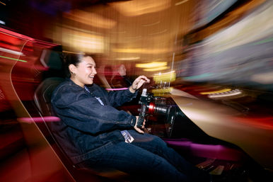 Smiling person in a denim jacket grips the wheel of an indoor arcade racing simulator, colorful motion-blurred lights streaking past and a second player visible in the background.