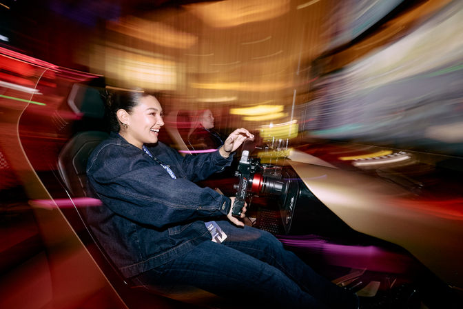 Smiling person in a denim jacket grips the wheel of an indoor arcade racing simulator, colorful motion-blurred lights streaking past and a second player visible in the background.