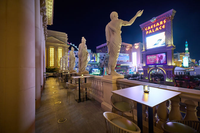 Roman-style marble statues on a balcony overlooking the neon-lit Las Vegas Strip at night with outdoor dining tables and glowing hotel signage