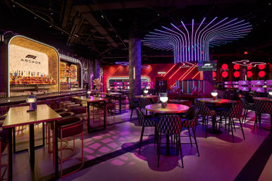 Neon-lit racing-themed arcade lounge with a backlit bar, high tables, round booths, rows of gaming stations and dramatic purple-pink LED ceiling artwork