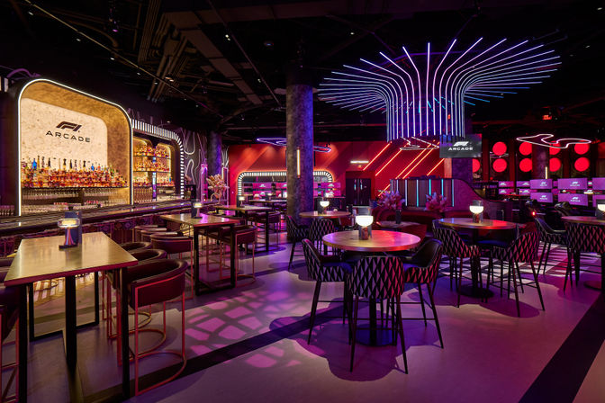 Neon-lit racing-themed arcade lounge with a backlit bar, high tables, round booths, rows of gaming stations and dramatic purple-pink LED ceiling artwork